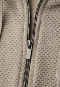 Primer plano de una prenda de tejido texturizado beige parcialmente cerrada con una cremallera de metal plateado.