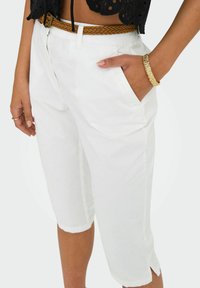 Bijele skraćene hlače ravnog kroja, s bočnim džepovima i prevrnutim rubom. Uparene s pletenim smeškim remenom i zlatnom narukvicom.