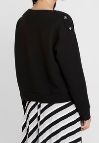 Svart sweatshirt med en avslappnad passform, ribbad textur och knappdetaljer på axeln. Bärs över en svart- och vitrandig kjol.