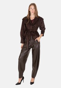 Giacca avvolgente in camoscio marrone con ampi revers, legata in vita. Abbinata a pantaloni in pelle marrone scuro con vestibilità comoda. Tacchi alti neri.