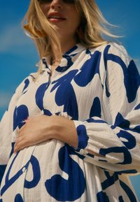 Witte blousejurk met opvallende blauwe abstracte patronen, textuurstof en lange mouwen met geplooide manchetten, gedragen over een ronde buik.