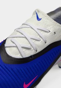 Αθλητικά παπούτσια με λευκό υφασμάτινο επάνω μέρος, μπλε κάτω τμήμα με αυλακώσεις και ροζ λογότυπο Nike. Ο σχεδιασμός περιλαμβάνει λευκά κορδόνια και μαύρες πινελιές.