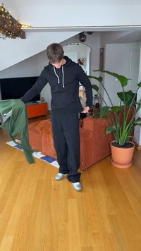 Černá mikina s kapucí a bílými šňůrkami, v ruce zelená látka, černé kalhoty a stříbrné boty. Vnitřní prostor s květináčem a nábytkem.