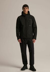 Czarna kurtka softshell z wysokim kołnierzem, zapinaną na zamek kieszenią na piersi oraz kieszeniami bocznymi. Noszona z dopasowanymi czarnymi spodniami i mocnymi czarnymi butami.