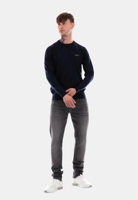Maglione a maniche lunghe di colore blu navy realizzato in tessuto a maglia, abbinato a jeans grigi. La persona indossa sneakers bianche e ha un tatuaggio sul collo.