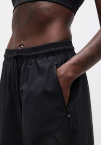 Κοντινό πλάνο ενός ατόμου που φοράει μαύρα αθλητικά σορτς με φερμουάρ στην τσέπη, με το χέρι να ακουμπάει μέσα στην τσέπη και εμφανές σκουλαρίκι στον αφαλό.