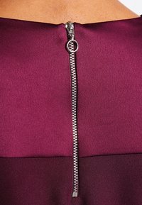 Mörkröd klänning med en mjuk tygtextur har en silverfärgad dragkedja på baksidan som sträcker sig vertikalt ner i mitten.