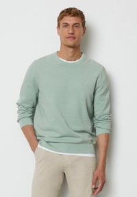Hellgrüner Strickpullover mit gerippten Bündchen und Saum, Rundhalsausschnitt und kleinem Logodetail auf der Brust. Über einem weißen Hemd getragen.