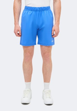 Homme portant un short de sport bleu vif, des chaussettes blanches et des baskets blanches, debout devant un fond clair uni.