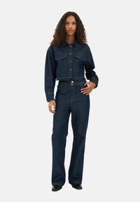 Sötét farmerből készült, gombos elülső részes, rövidített dzseki, két mellzsebbel, magas derekú, széles szárú sötét farmernadrággal párosítva. Sima textúra.