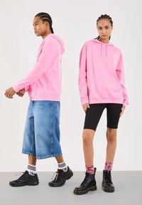 Zwei Models mit geflochtenen Haaren tragen rosa Kapuzenpullover; die eine kombiniert ihn mit weiten Denim-Shorts und Sneakern, die andere mit schwarzen Radlerhosen und Stiefeln.