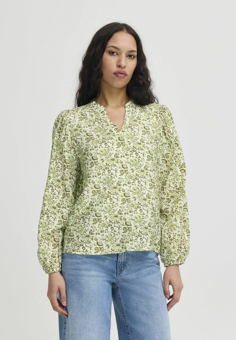 Blusa verde con stampa floreale, scollo a V, maniche a sbuffo e dettagli arricciati. Realizzata in tessuto leggero, abbinata a jeans blu dal taglio rilassato.