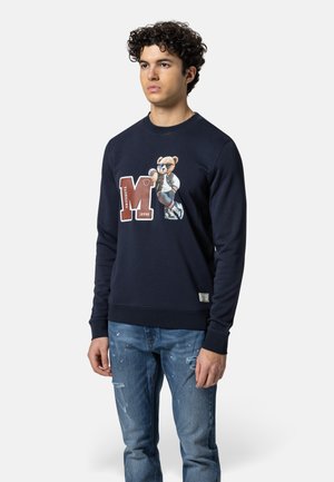 Junger Mann trägt ein marineblaues Sweatshirt mit einem großen "M" und einem Bär-Motiv, kombiniert mit abgenutzten blauen Jeans.