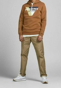 Felpa marrone con stampa grafica, pantaloni beige e sneakers bianche. La felpa presenta un cordino e polsini a costine.