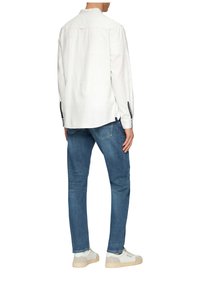Witte langemouwen shirt met een kraag, zijsplitten en marineblauwe accenten op de mouwen, gecombineerd met blauwe jeans en witte sneakers.