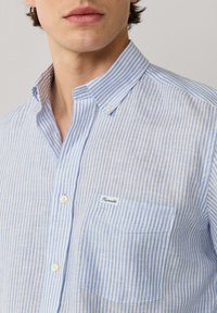 Hombre vistiendo una camisa abotonada de rayas verticales en azul claro y blanco, con cuello abotonado y un pequeño bolsillo en el pecho con la etiqueta "Façonnable".