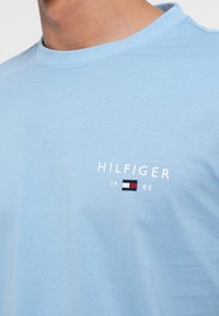 Tricou din bumbac de culoare albastru deschis, cu guler rotund. Prezintă un text brodat "HILFIGER" și un mic logo în culorile roșu, alb și albastru. Textură netedă.