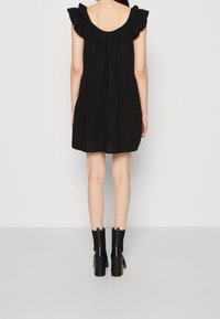 Vestido negro corto con mangas rizadas, diseño de capas y escote redondeado en la espalda. Usado con botas de tacón negras hasta el tobillo.