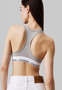 Sujetador deportivo gris con diseño de espalda nadadora, que presenta una banda blanca con el logo "Calvin Klein". Tejido suave con una silueta ajustada.