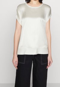 Haut en satin crème avec manches courtes retroussées, col rond et coupe légèrement décontractée. Associé à un pantalon noir à jambes larges avec coutures apparentes.