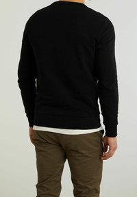 Svart långärmad tröja med ribbade manschetter och fåll, tillverkad av texturerad bomull, bärs över en vit skjorta och tillsammans med olivgröna byxor.