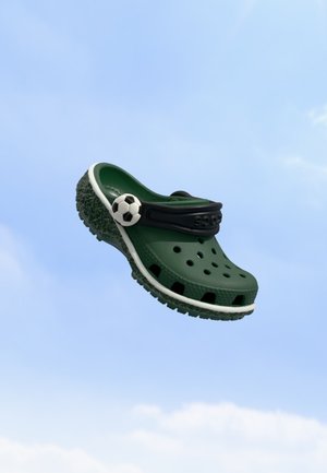 Un sabot vert foncé avec des trous d’aération, une bride noire au talon et un charm en forme de ballon de football flotte sur fond de ciel bleu.