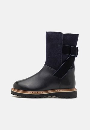 Bottine en cuir noir et daim avec surpiqûres marron, semelle épaisse, et sangle latérale décorative avec boucle.