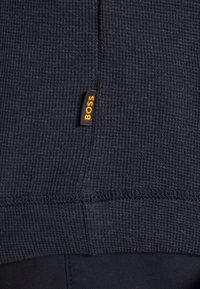 Gros plan sur un tissu noir texturé avec une petite étiquette noire portant l'inscription "BOSS" en lettres orange cousue le long d'une couture.