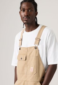 Άνδρας με σκούρα πλεξούδες φοράει μπεζ ολόσωμη φόρμα πάνω από λευκή μπλούζα, κοιτάζοντας εμπρός με φόντο ένα απλό ανοιχτόχρωμο τοπίο.