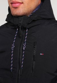 Giacca nera imbottita con colletto alto, zip frontali e lacci di contrasto blu, rossi e bianchi; presenta il logo sul petto.