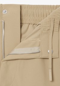 Gros plan sur la ceinture d'un pantalon beige avec un dos élastique, une fermeture éclair partiellement ouverte, un bouton pression et un cordon de serrage avec une étiquette de marque.