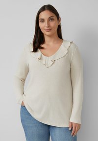 Hellbeige geripptes Langarmoberteil mit V-Ausschnitt und Rüschendetail am Kragen. Kombination mit blauen Jeans in entspannter Passform.