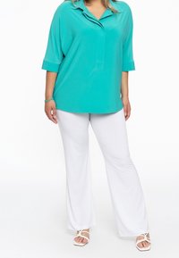 Blouse turquoise avec un col et des manches trois-quarts, associée à un pantalon blanc évasé et des sandales blanches à lanières.