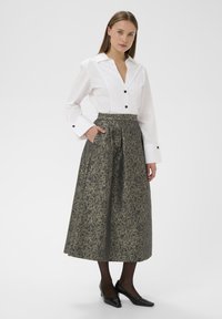 Balta marškiniai su juodais mygtukais ir rankovėmis derinami su ilga, tekstūruota, tamsiai žalia ir auksine A formos sijonu, turinčiu šonines kišenes. Juodi aukšti kulniukai.