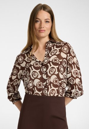 Frau mit langem hellbraunem Haar, die eine braune und beige gemusterte Bluse und einen dunkelbraunen Rock vor einem schlichten weißen Hintergrund trägt.