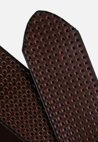 Tira de chancla marrón texturizada con un patrón de diamantes y bordes lisos, con un borde negro de contraste. Material de goma duradero.