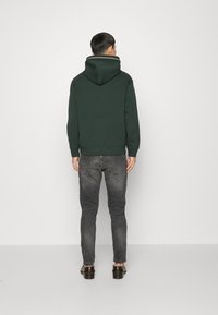 Grön hoodie med en stor huva, ribbade muddar och nederkant, bärs över grå denimjeans. Enkelt mönster, slät textur och inga synliga mönster.