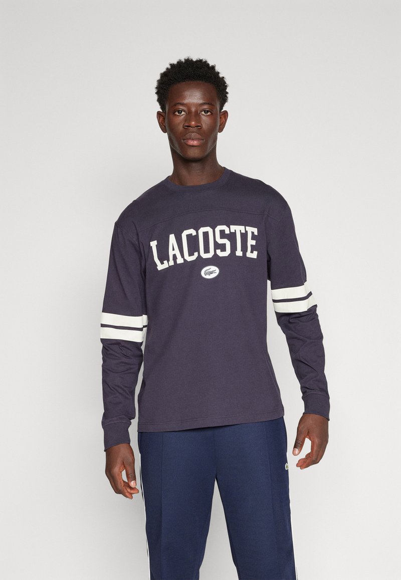 Jongeman met kort krullend haar, gekleed in een marineblauwe Lacoste longsleeve met witte strepen op de mouwen en marineblauwe broek, staand tegen een effen achtergrond.