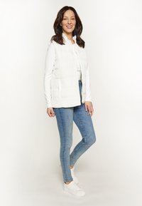 Weißer Steppwesten mit Reißverschluss, getragen über einem Langarmoberteil, kombiniert mit blauen Jeans und weißen Sneakers. Glatte Textur, lässige Passform.
