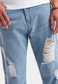 Primer plano de unos jeans rasgados de color azul claro, llevados por una persona con las manos reposando cerca de los bolsillos y hilos blancos visibles en las áreas desgastadas.