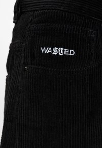 I pantaloni in corduroy neri presentano righe verticali, una tasca a toppa e una scritta ricamata bianca che recita "WASTED" sul retro.