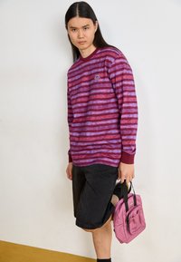 Chemise à manches longues rayée violet et rouge avec des poignets bordeaux, associée à un short noir et un sac à main rose avec une poche avant.