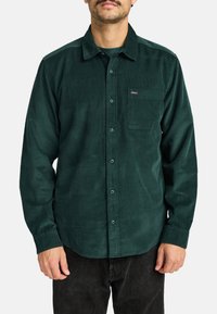 Green corduroy shirt with a pointed collar, button front, single chest pocket, and a textured fabric. Worn over dark jeans.