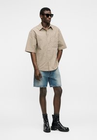 Hombre con gafas de sol negras, camisa de manga corta a cuadros beige, pantalones cortos de mezclilla y botines negros, de pie contra un fondo blanco.