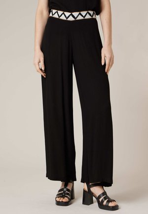 Femme portant un pantalon large noir avec une ceinture blanche et noire à motifs, ainsi que des sandales à talons hauts noires à brides.
