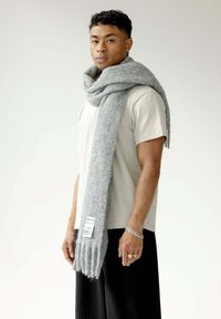 Écharpe grise et texturée en matériau moelleux, drapée autour du cou de manière décontractée, avec des extrémités à franges ; étiquette à une extrémité. Portée avec une chemise légère.