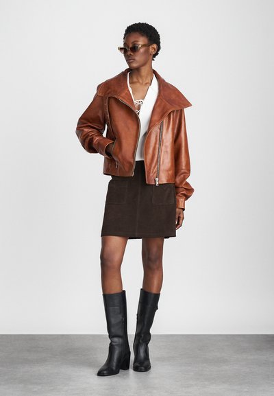Chaqueta de cuero marrón corta con cuello ancho, blusa blanca, mini falda de ante oscura con bolsillos delanteros y botas negras hasta la rodilla.