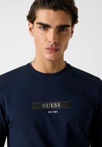 Granatowa koszulka z długim rękawem wykonana z bawełny, posiada prostokątną czarną naszywkę z wyhaftowanymi na biało napisami „GUESS” i „EST. 1981”. Gładka tekstura.