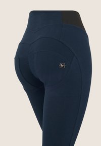 Primo piano di leggings a vita alta blu navy con cuciture curve e piccolo logo sulla tasca laterale.