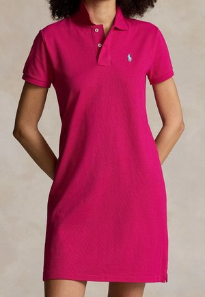 Femme portant une robe polo à manches courtes de couleur rose vif avec un petit logo brodé bleu sur la poitrine, debout les mains derrière le dos.